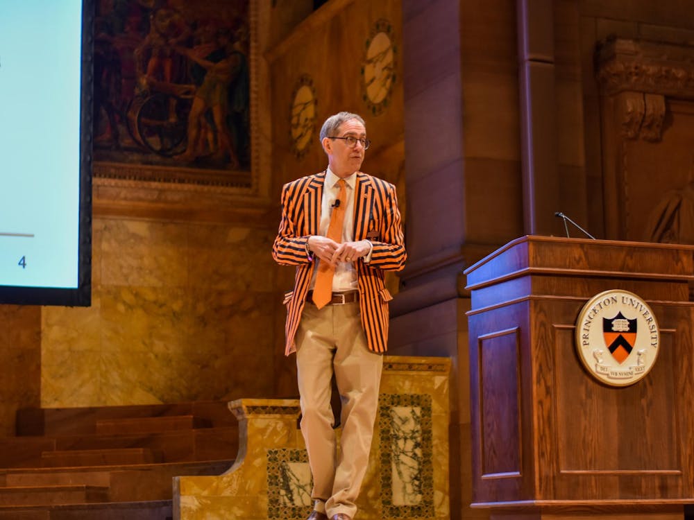 A man in a black and orange striped blazer and orange tie stands next to a podium adorned with a Princeton University crest.