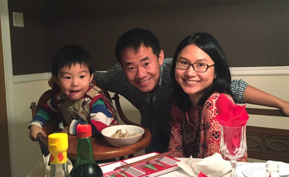 Asian man stands behind Asian woman and toddler seated. There is an empty glass in the frame and the toddler is in a high chair eating dinner.