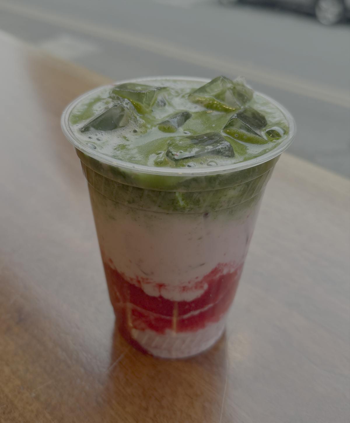 An iced matcha latte in a clear plastic cup sits on a wooden table, with red syrup in the center. 