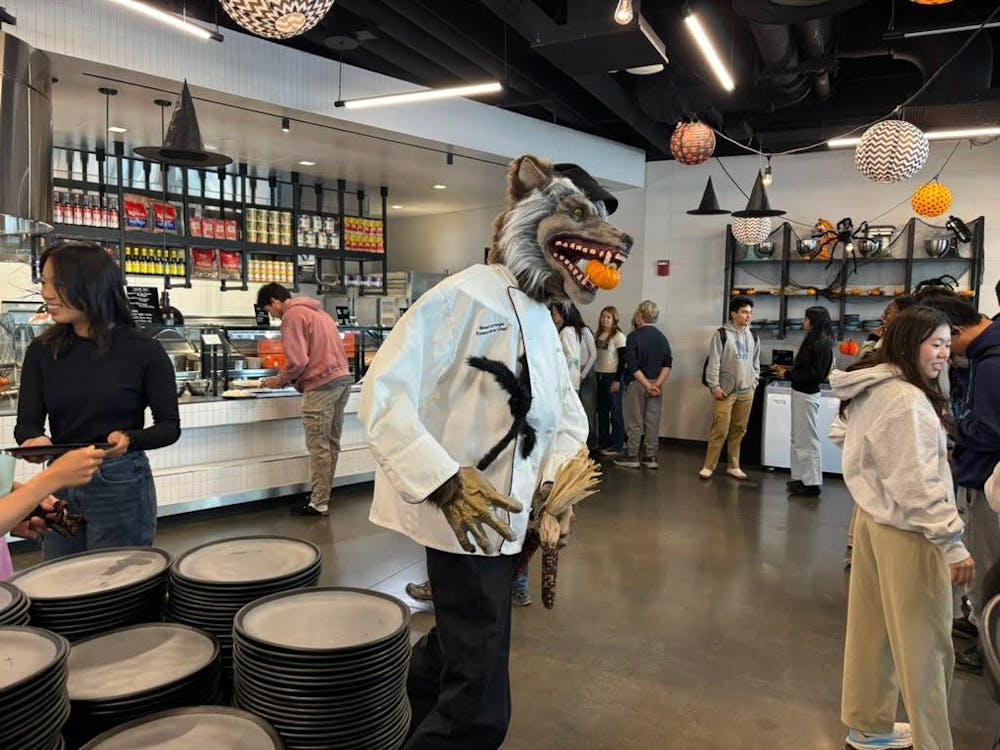 NCW dining hall decorated in Halloween decor. Several students in shot. In the center is a werewolf figure, dressed in a white campus dining shirt, with a miniature pumpkin in its mouth.