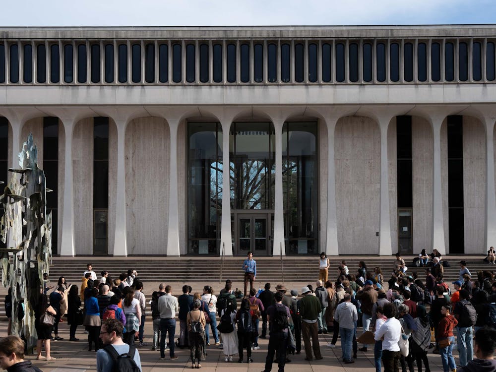 A large group of people stand outside a modern white building with columns. A fountain is to the left.