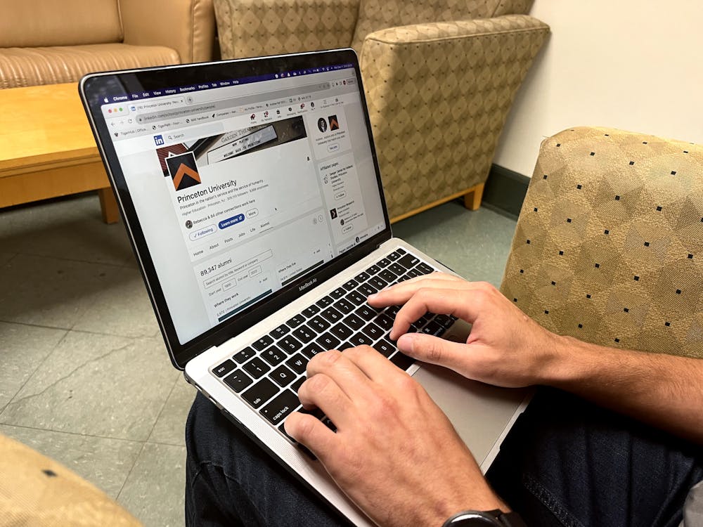 Hands of a student on a grey computer keyboard with a computer screen displaying the Princeton University LinkedIn page, with chairs in the background