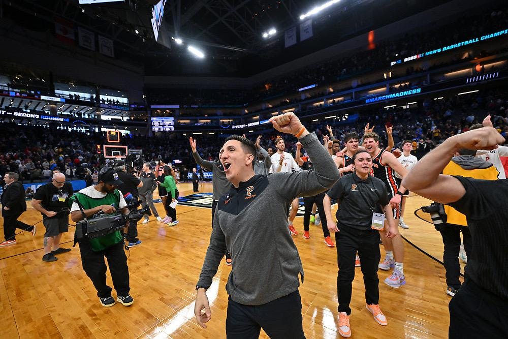 A Princeton basketball staff member celebrates on the court following the Tigers’ NCAA Tournament upset, with players and arena signage visible in the background. 