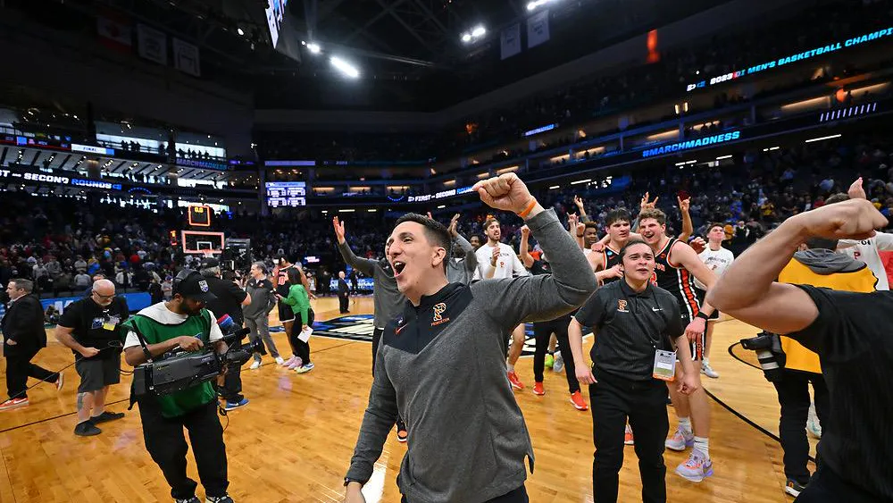A Princeton basketball staff member celebrates on the court following the Tigers’ NCAA Tournament upset, with players and arena signage visible in the background.