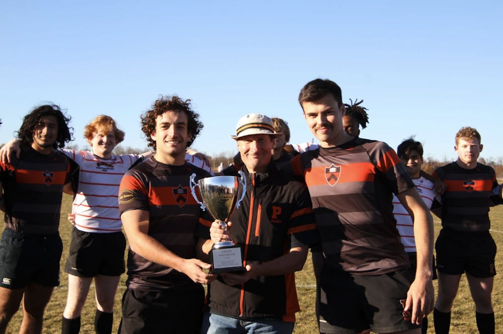 Coach Richard Lopacki holding the alumni game trophy, standing alongside two Class of 2024 men's rugby players.