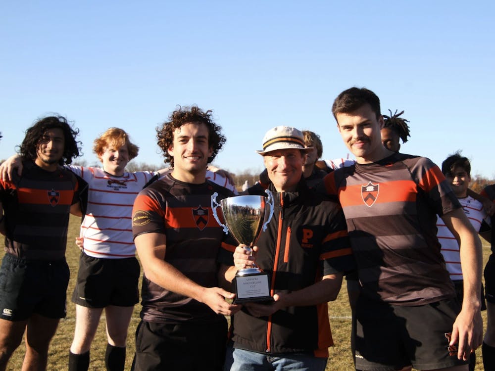 Coach Richard Lopacki holding the alumni game trophy, standing alongside two Class of 2024 men's rugby players.