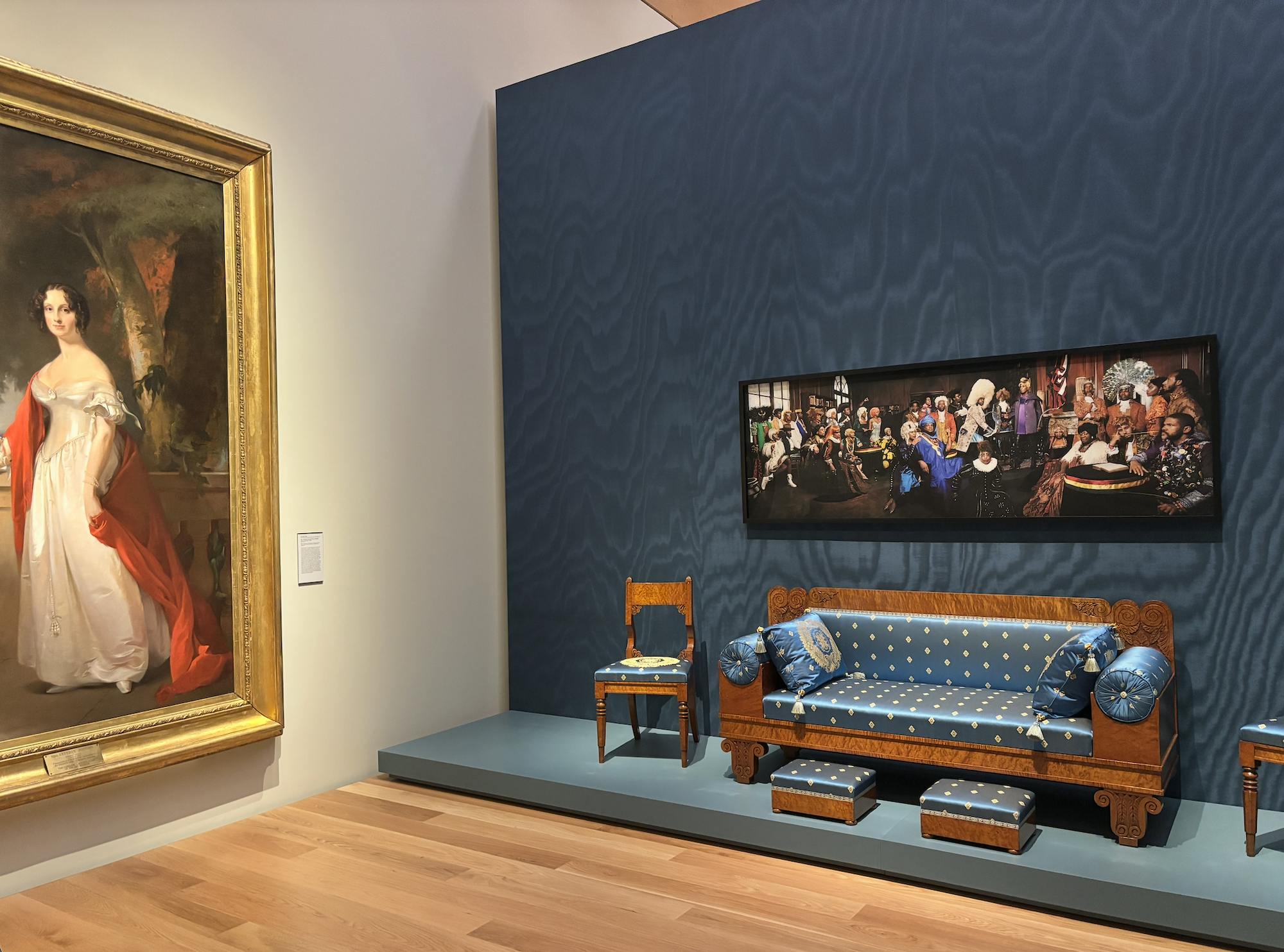 A long horizontal painting depicts people of color, wearing multicolor clothing, in the signing of the constitution. The painting hangs on navy blue textured wall. Under the painting is a blue couch with white dots, with a brown wood back.