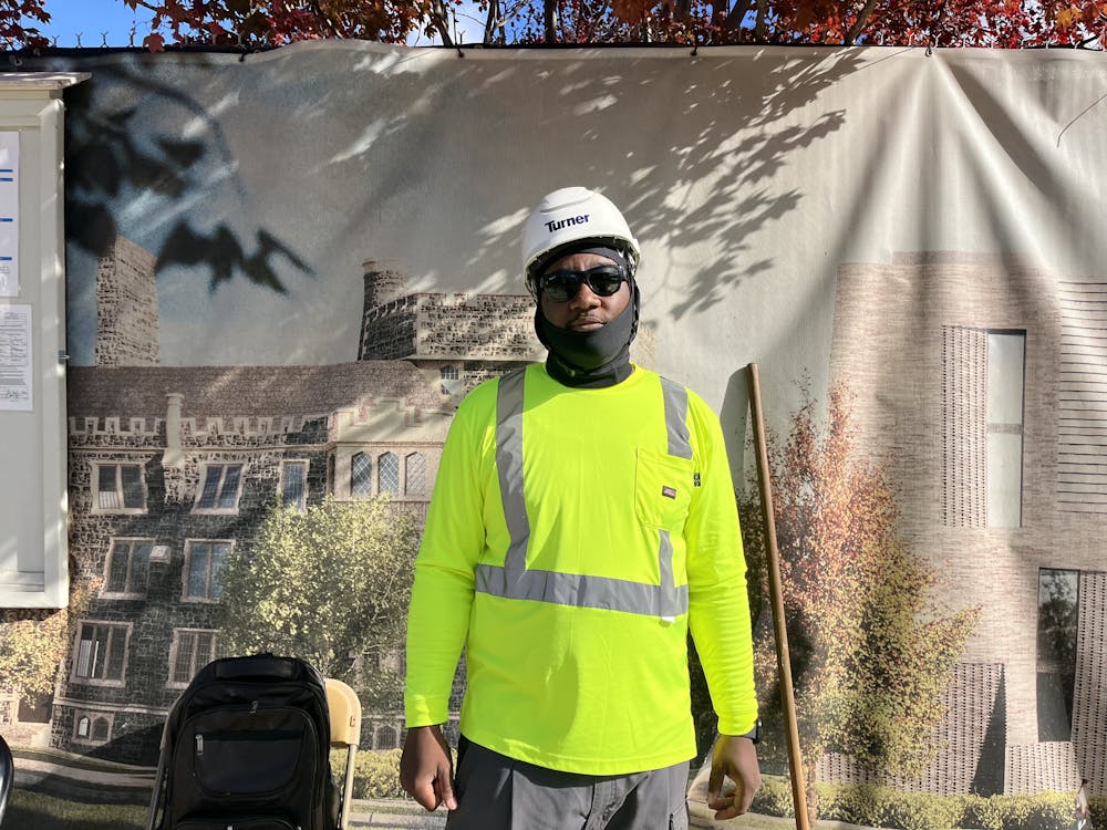 A man in a construction hat, sunglasses, and neon vest is photographed in front of a construction site. 