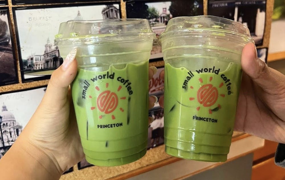 Two hands each holding a green oat-infused matcha in clear cups from Small World. The cups display the circular red Small World logo surrounded by the words "Small World Coffee Princeton". The matcha drinks are in front of a wall adorned with photographs. 