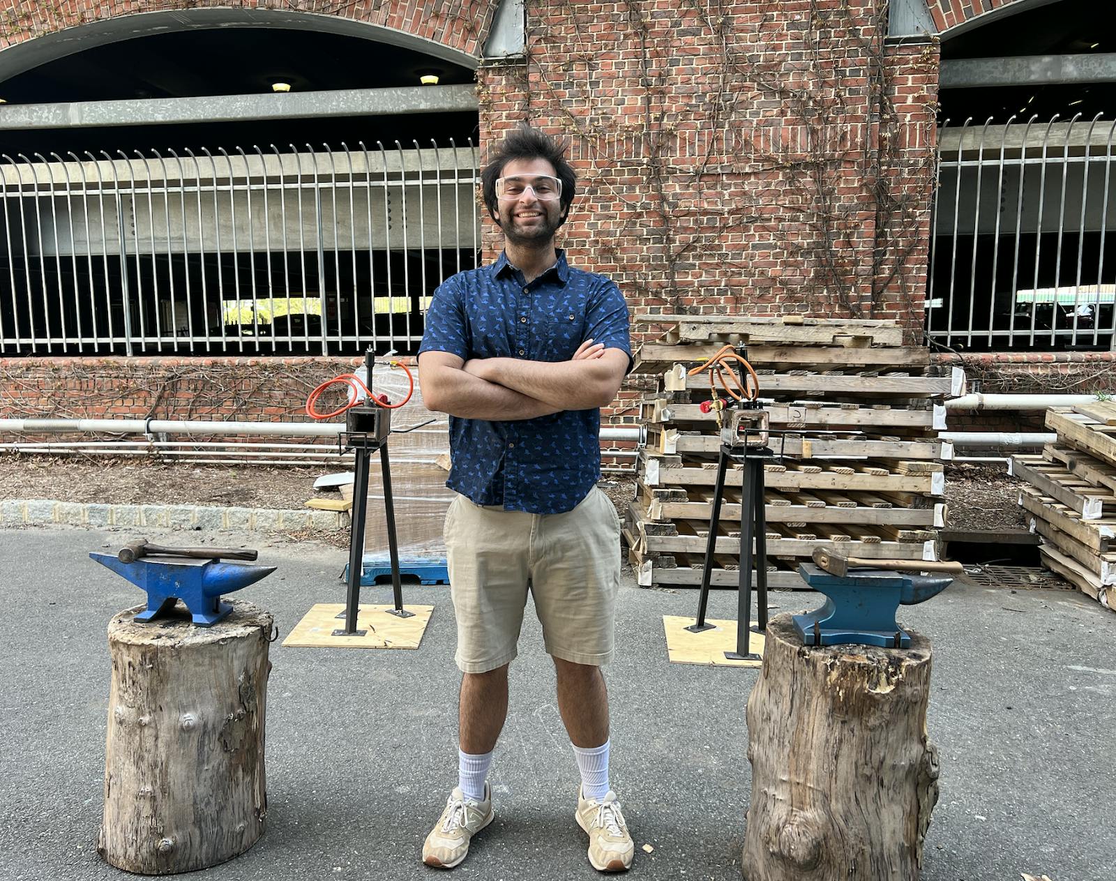 A young male student with his arms crossed in front of him and smiling, standing in front of some anvils and forge materials in an alleway.