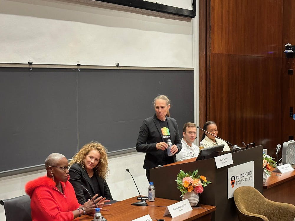 Five speakers are at a podium, with the person in the middle, a woman with a gray ponytail, standing. A chalkboard is in the background.