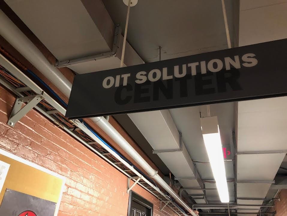 A gray sign hangs from the ceiling reading “OIT Solutions Center” and the wall is red brick.
