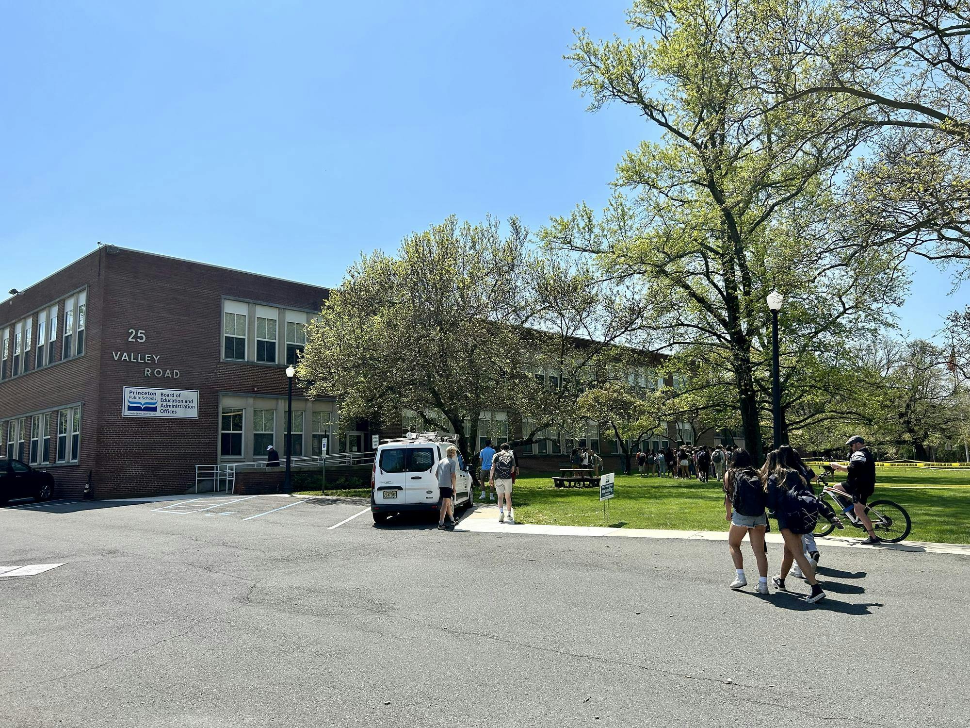 Students wearing backpacks walk across a parking lot toward a brown school building.