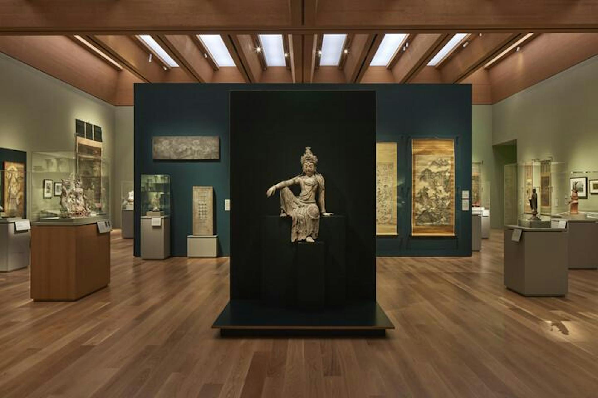 A sculpture is raised in front of a dark wall. There is a room surrounding the sculpture containing other pieces of art.