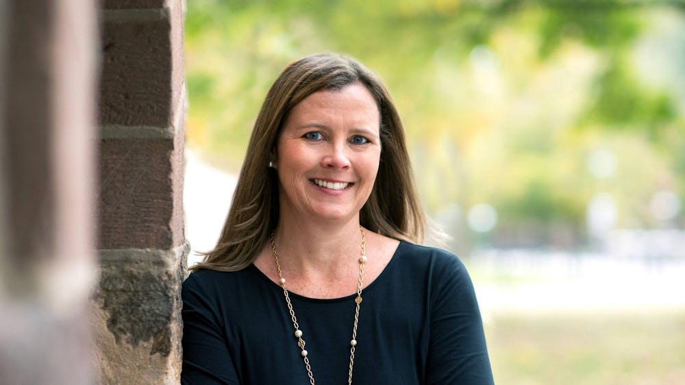 A woman with brown hair, a gold necklace, and a black top leans against a brick wall.