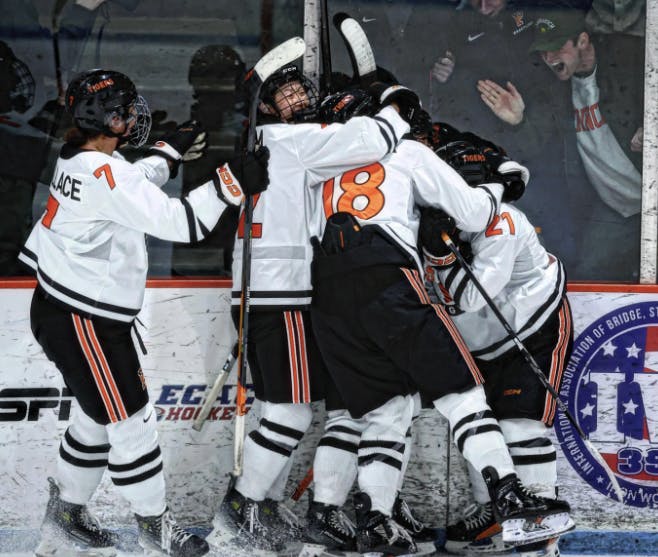 Players in white and orange uniforms hug another player with fans cheering through the glass. 