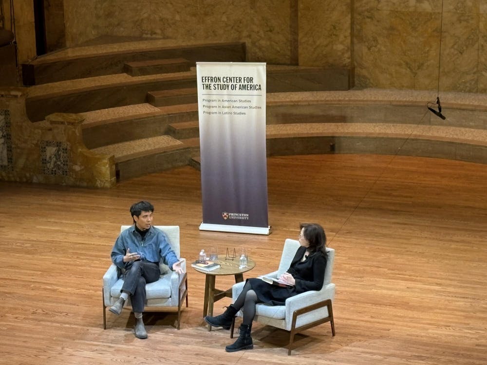 A man in a stylish blue jacket with light brown boots sits with a woman dressed in all black on a big wooden stage. 