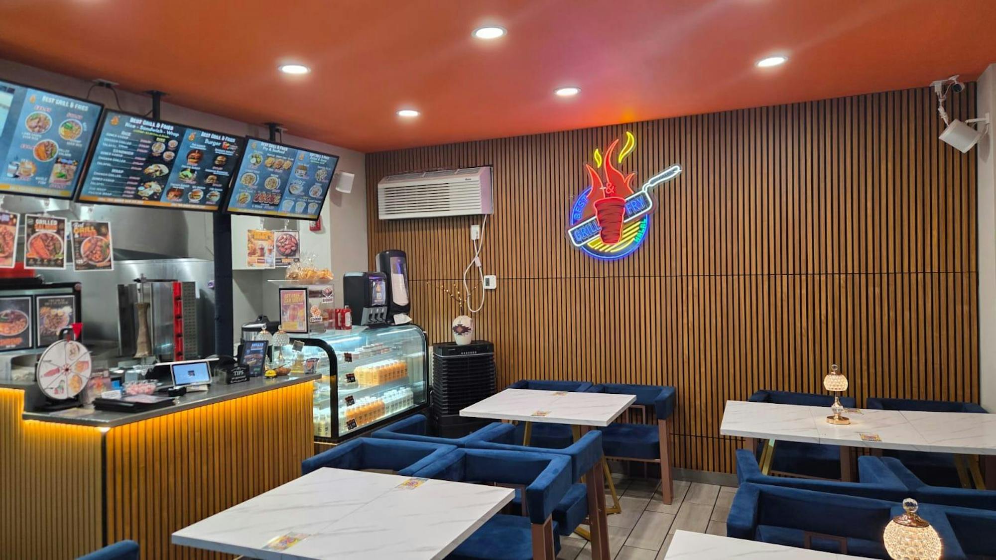 There is a small room with wood panels and several white tables and blue cushioned chairs. There is a counter on one side of the room with TV menus and a glass case with food items in it.