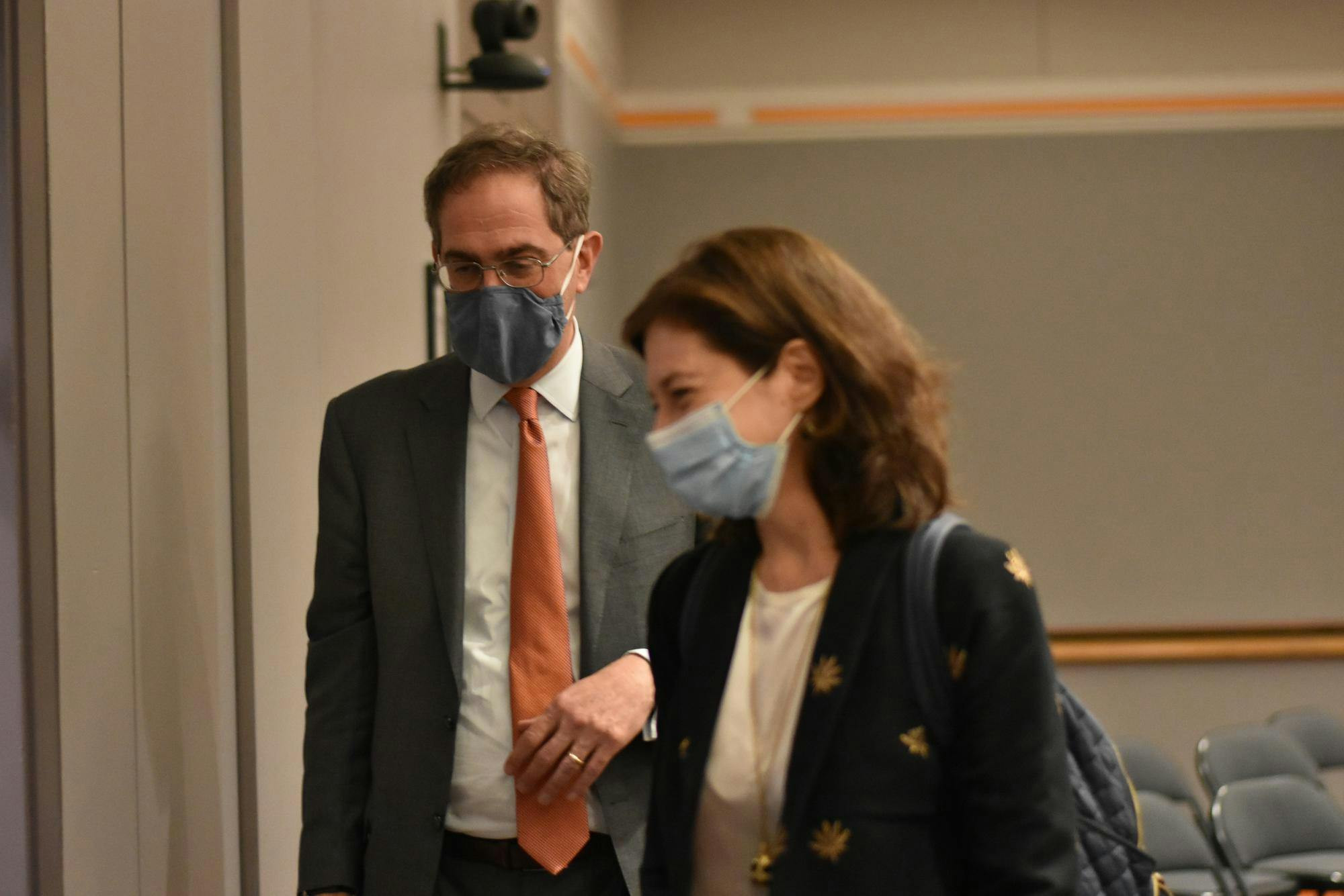 A man wearing a face mask looks down, slightly behind a woman wearing a face mask.