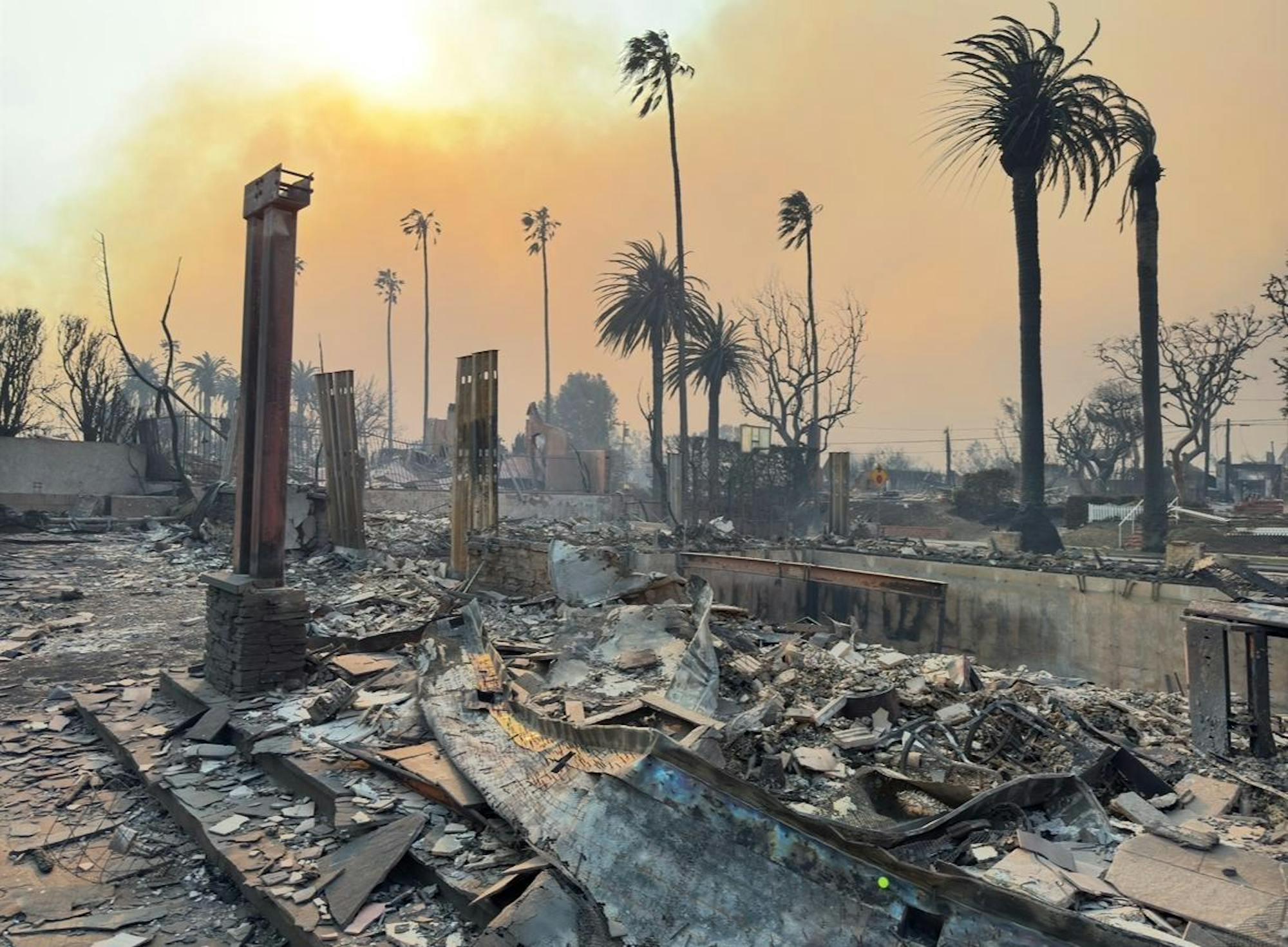 A few tree trunks stand amidst rubble, in front of a sky that is orange as a result of the LA wildfires.