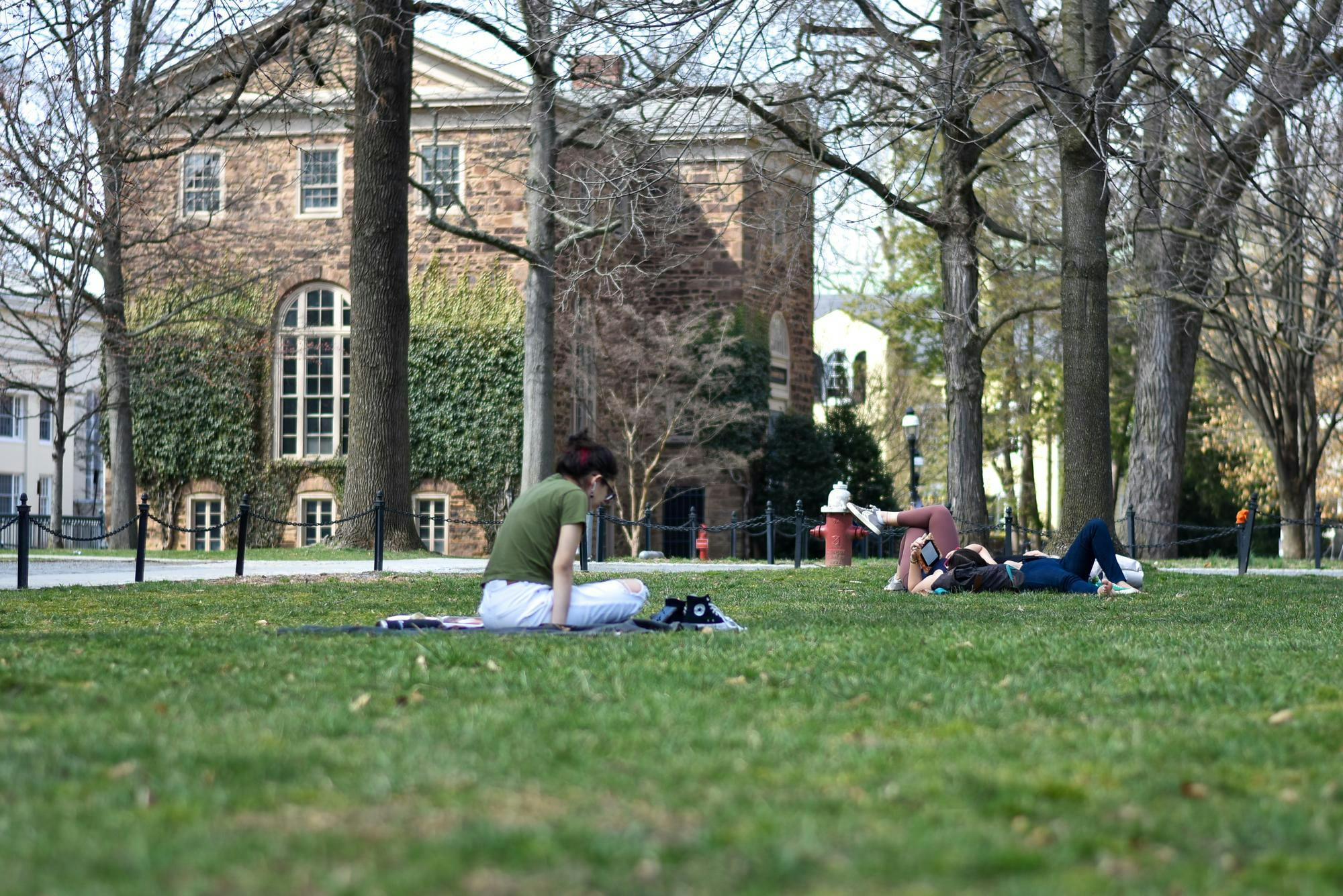 Two people sit on grass lawn with a brown stone building in the background.