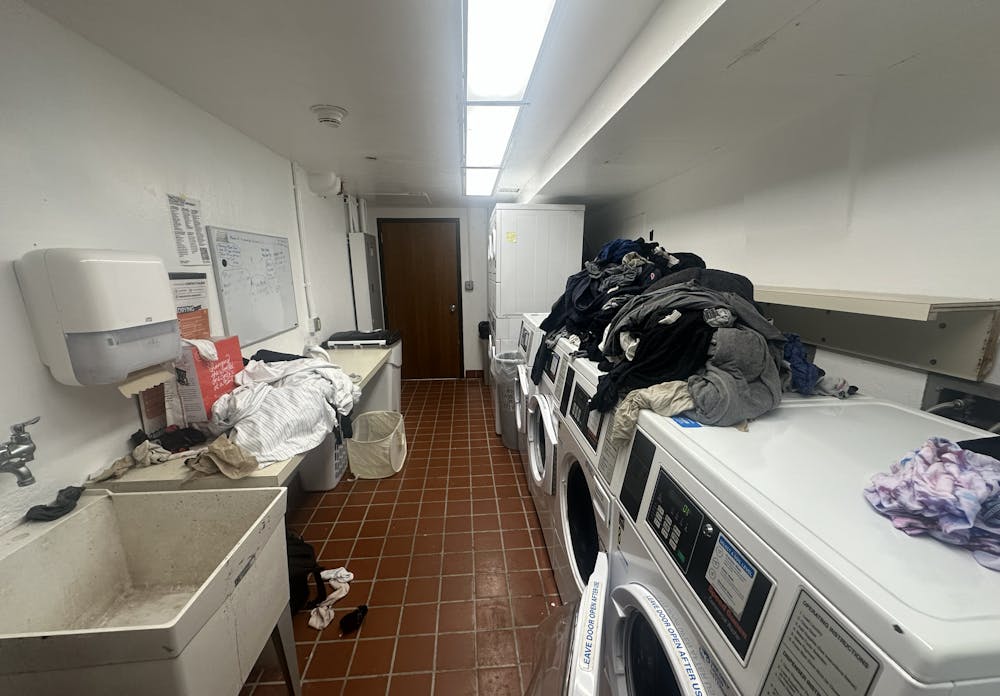 Laundry room with a row of washers and dryers. Clothes are piled over the washers. The room also contains a sink and a table.