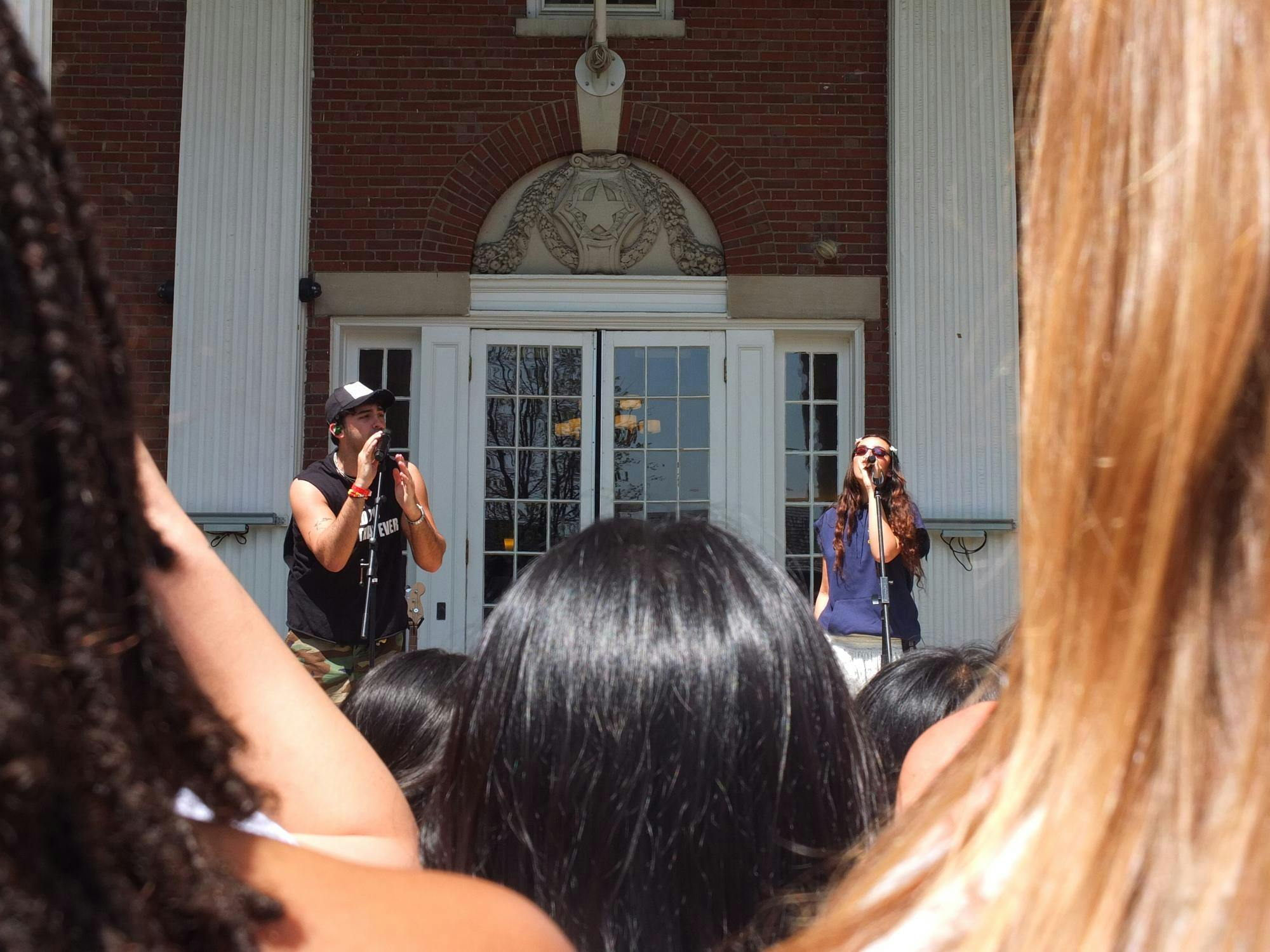 Two performers sing into microphones outside of a brick building with Ionic columns. 