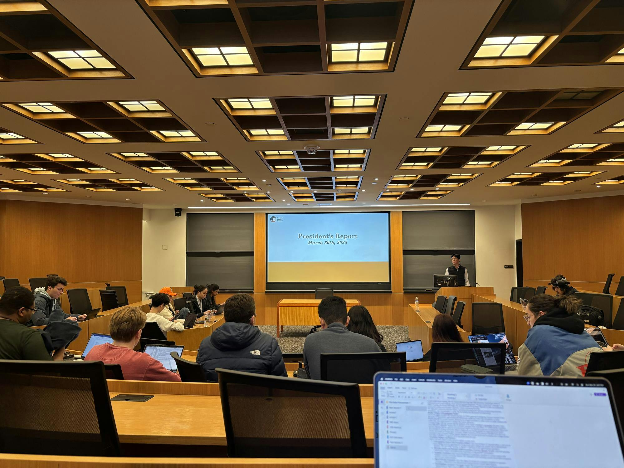 A number of people sit around a room. There is a large screen in the background which reads "President's Report, March 30th, 2025."