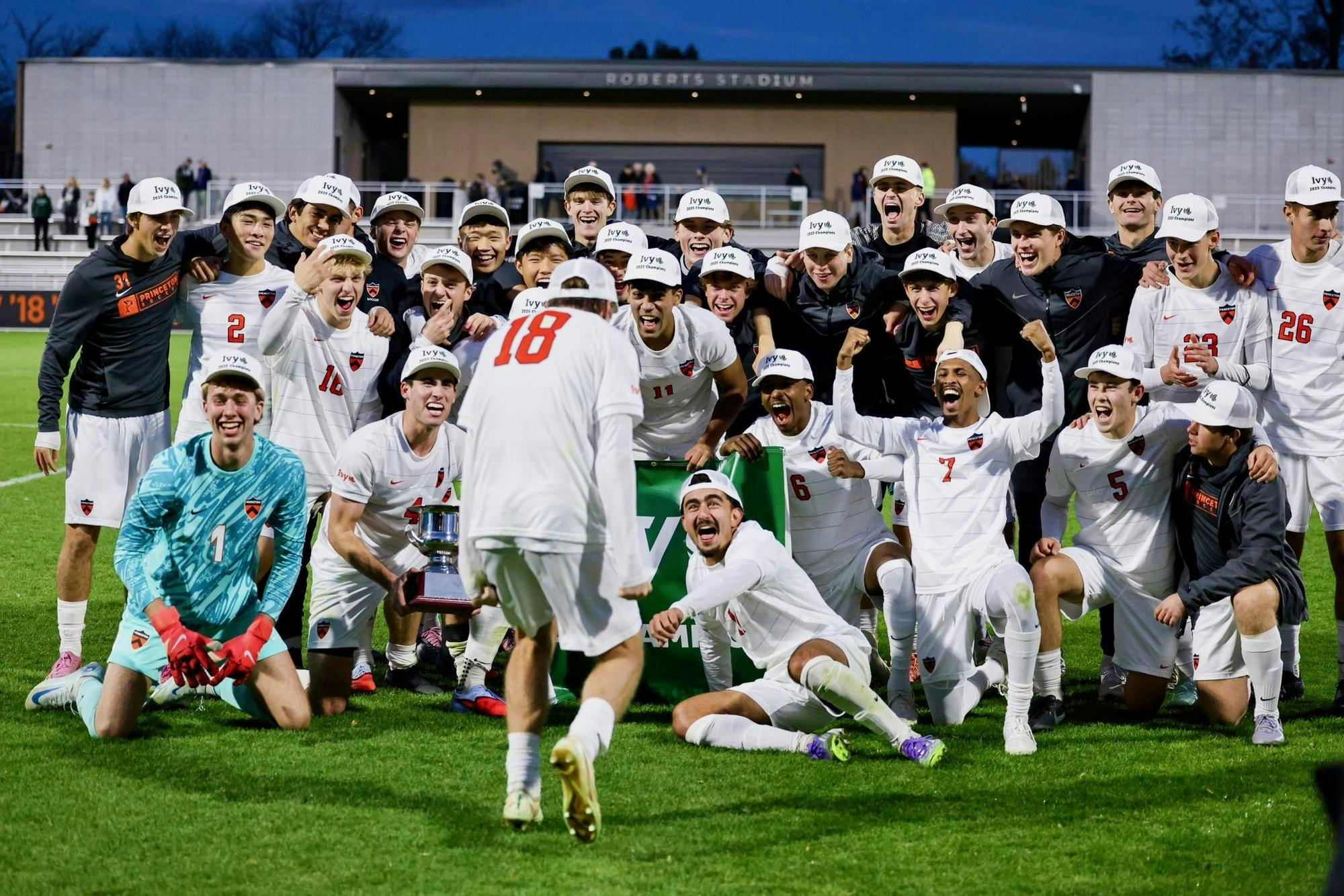 A group of soccer athletes wearing white shorts, white jerseys, and customized hats, celebrating a title on a grass field.