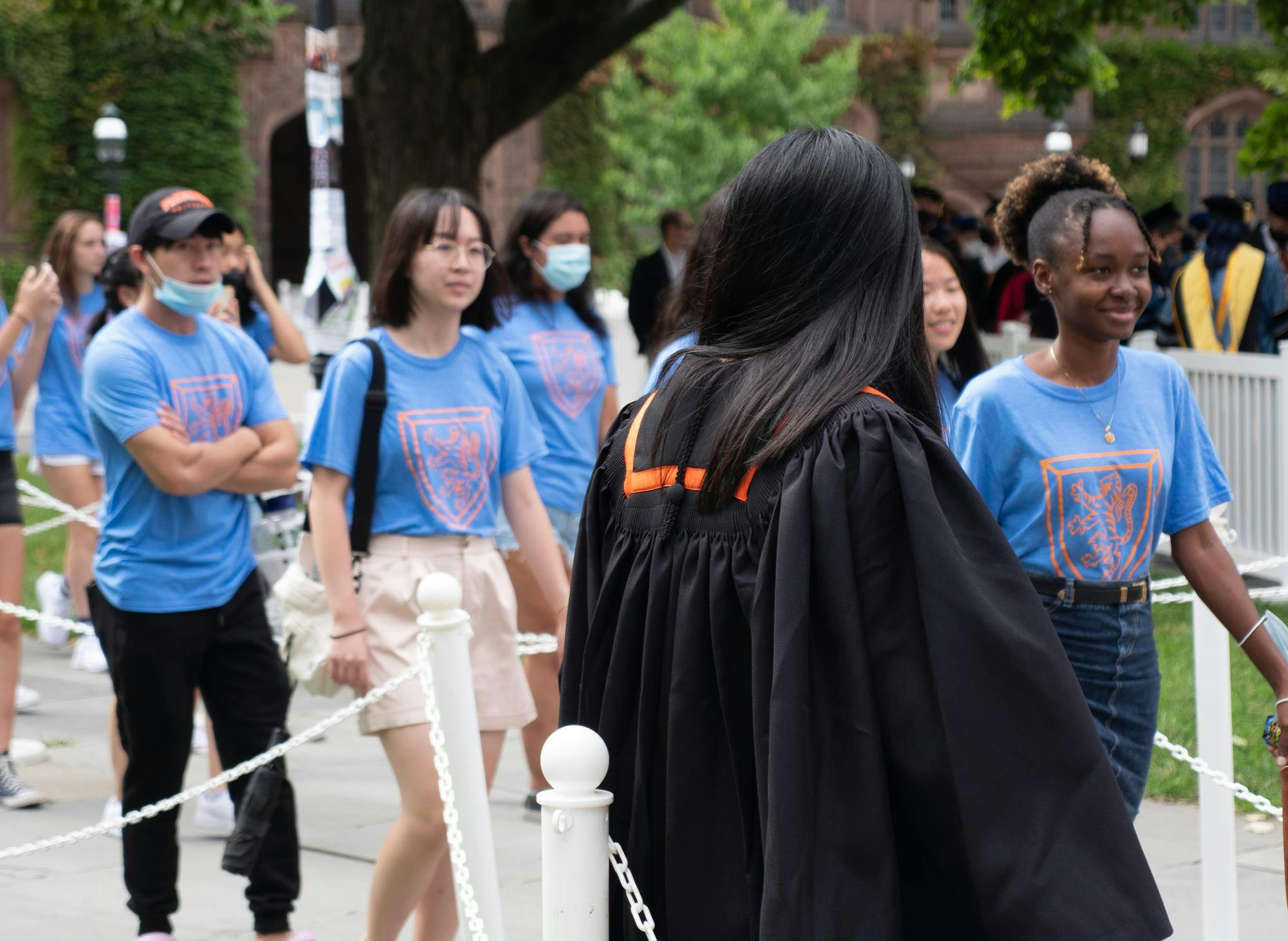 Students in light blue t shirts walk in a line outside a brown stone building.