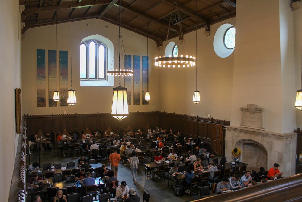 A large group of students sits in a large dining hall with high ceilings and large windows. 