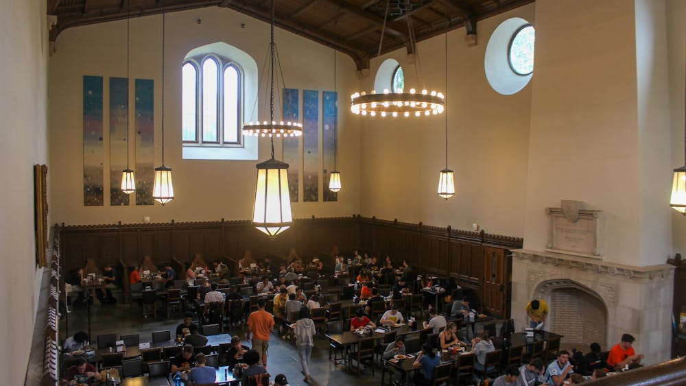 A large group of students sits in a large dining hall with high ceilings and large windows. 