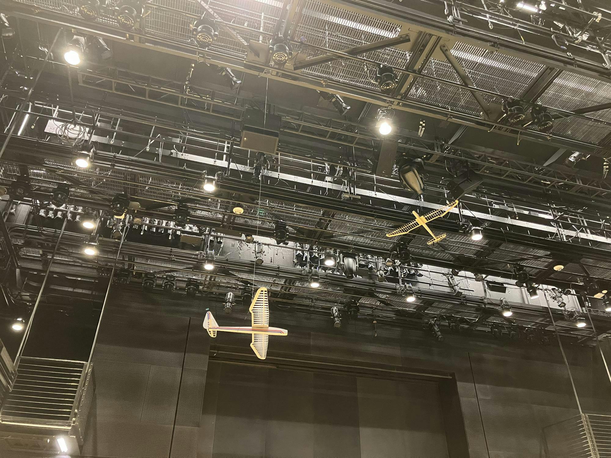 Two wooden airplanes are hung from a black theatre ceiling.