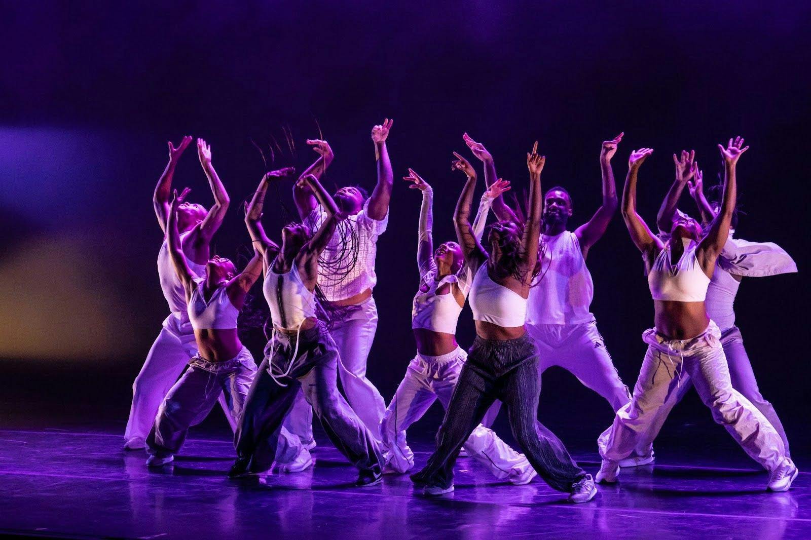 A group of nine dancers dressed in jeans and white tops mid-movement. The lighting gives the dancers a purple hue.