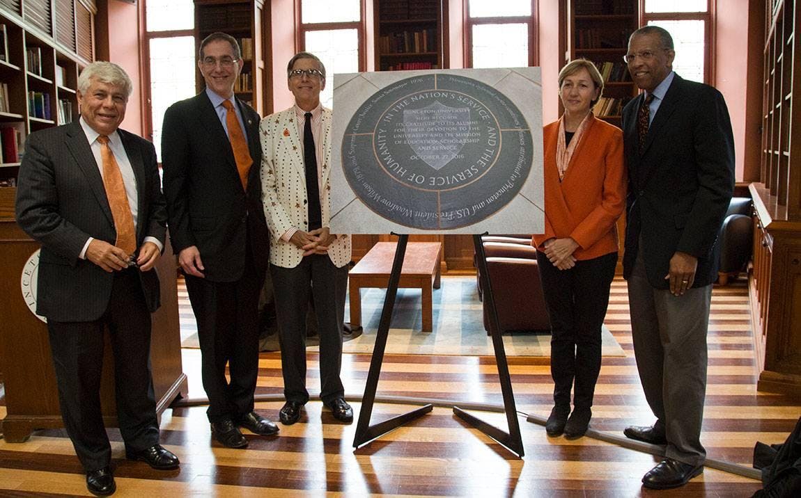 Five people stand behind an image of the revised informal motto mounted on a tripod. 