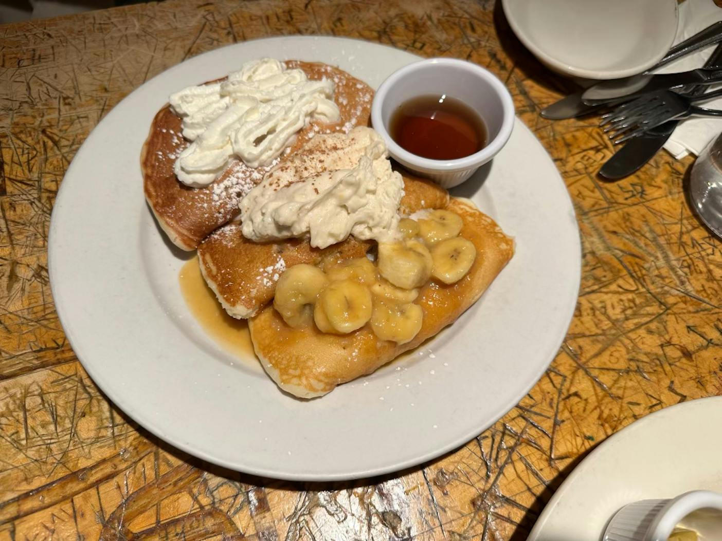 A white plate sits on a light brown table that is covered in small writings, scratches and pencil marks. There are three brown pancakes on the plate. There are three toppings: white cream, white cream with cinnamon, and banana pieces. There is also a small white container of brown syrup.