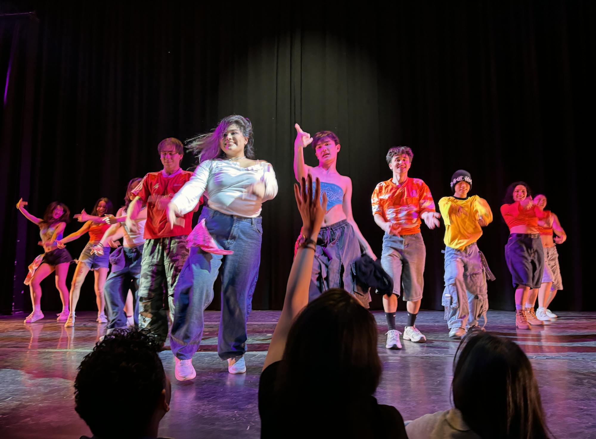 Several dancers stand in a v-shape on a black stage. They are all wearing different colors of street-style outfits.