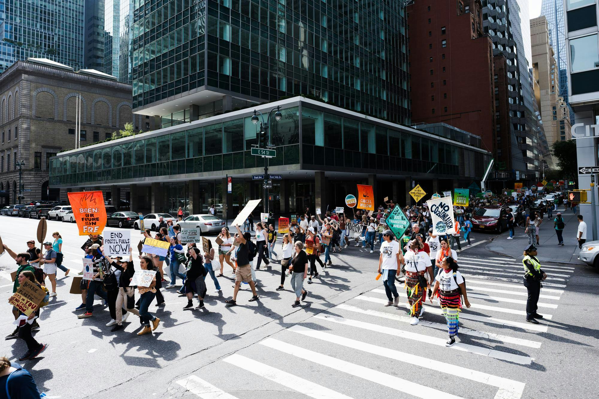 People walking and protesting with signs on a city street, with buildings in the background.