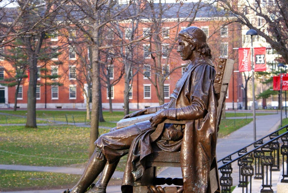 A bronze statue of a man sitting on a chair sits in front of a courtyard with trees and buildings.