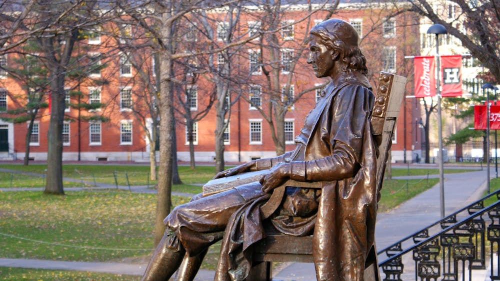 A bronze statue of a man sitting on a chair sits in front of a courtyard with trees and buildings.