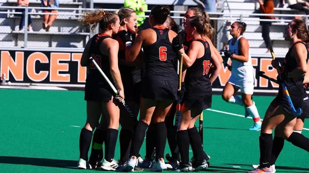 Women in black and orange uniforms huddle on a green field. 