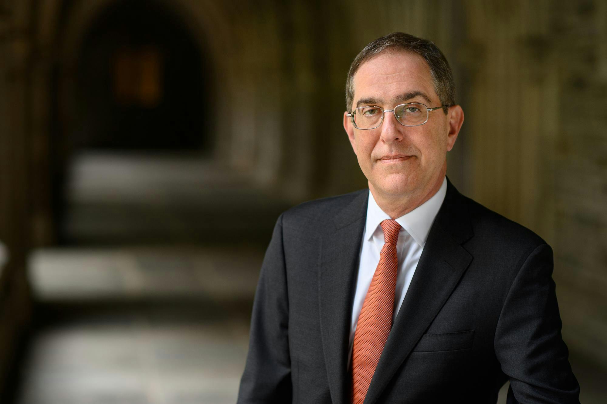 A man wearing a gray suit, white shirt, orange tie, and glasses with a blurred background.