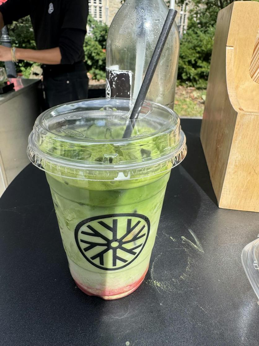 An iced matcha latte in a plastic cup sits on a black table by the Ooika stand. 