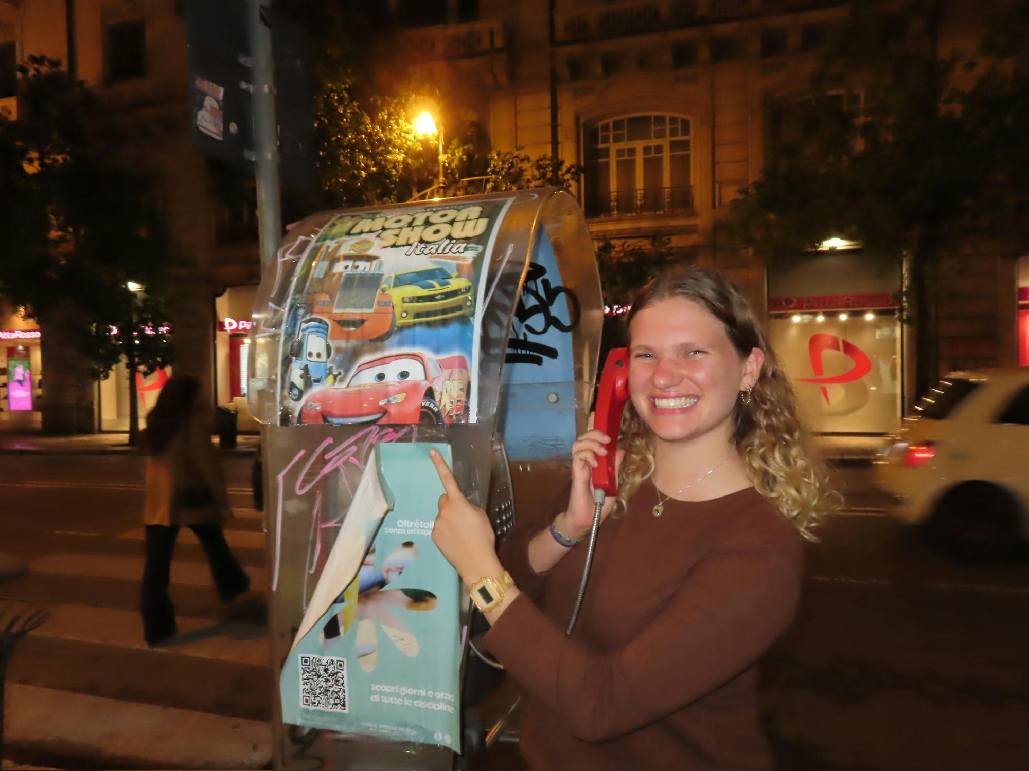 A young woman with blond hair and a brown shirt standing next to a silver telephone booth with a red lightning McQueen car poster on it.