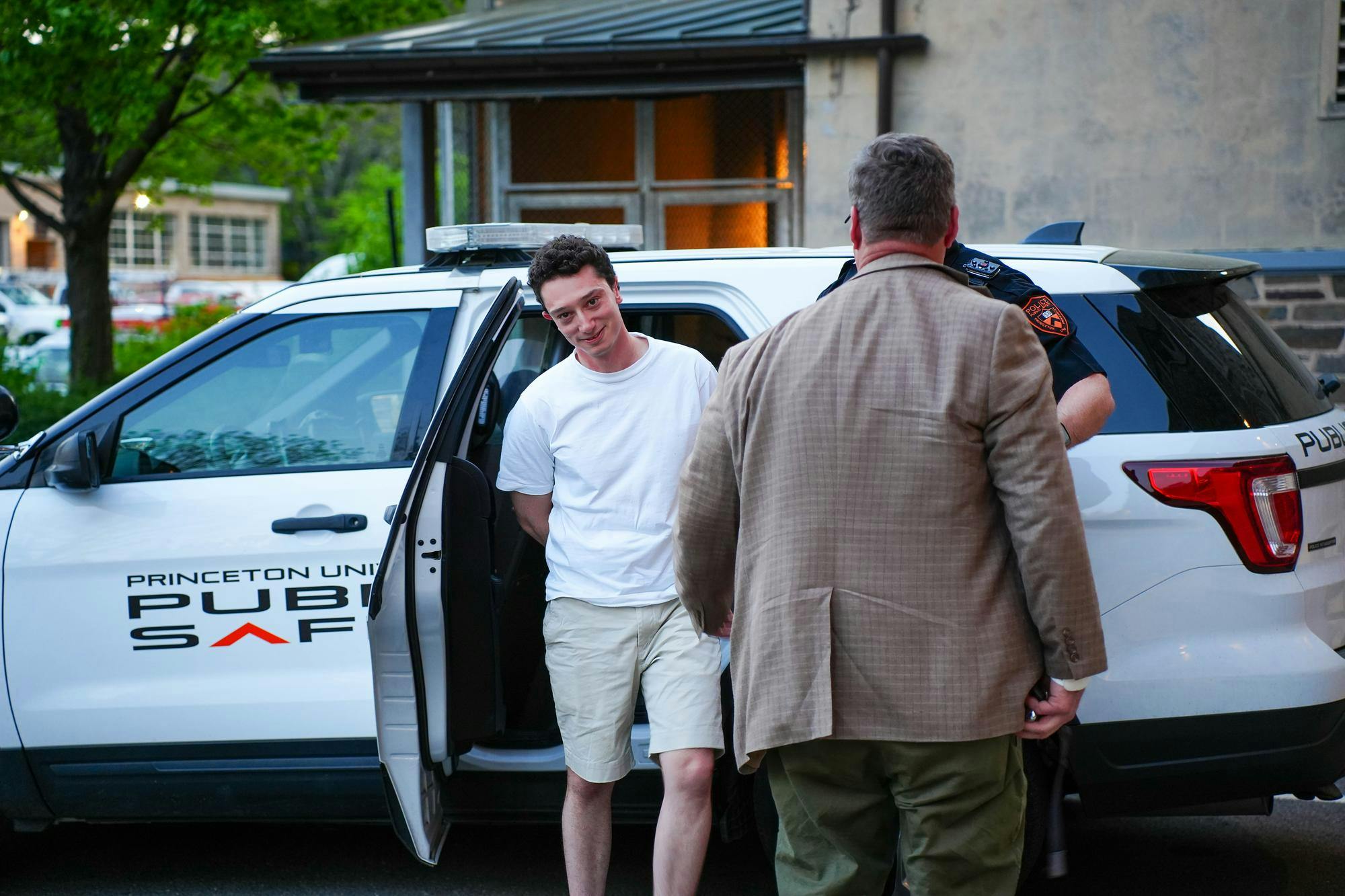 Man in a white shirt looks into the camera with his arms behind his back. Behind him, a car that reads “Princeton Public Safety”