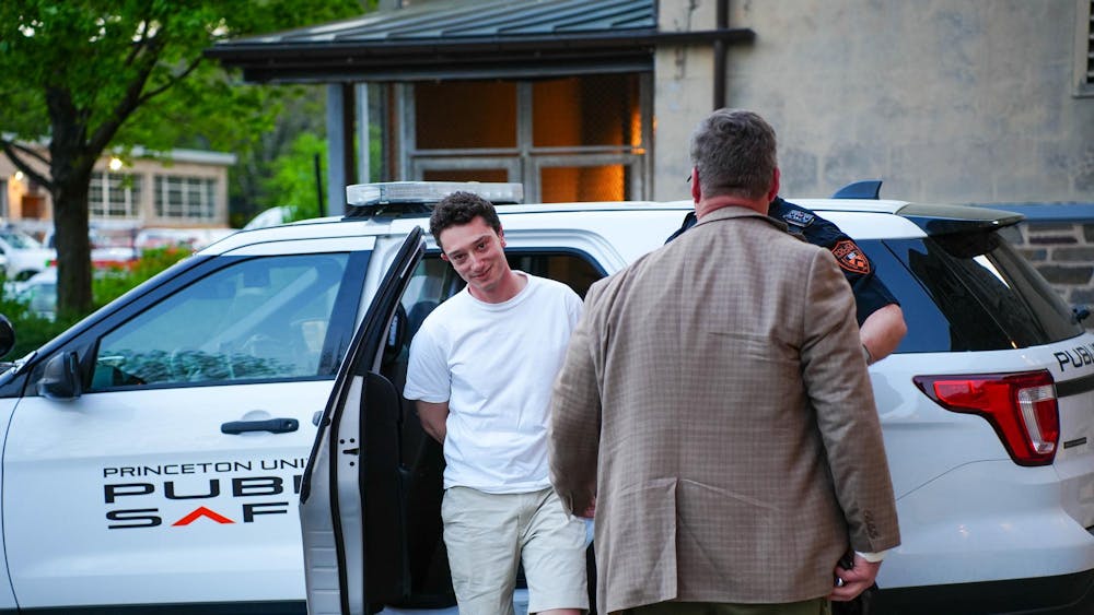 Man in a white shirt looks into the camera with his arms behind his back. Behind him, a car that reads “Princeton Public Safety”