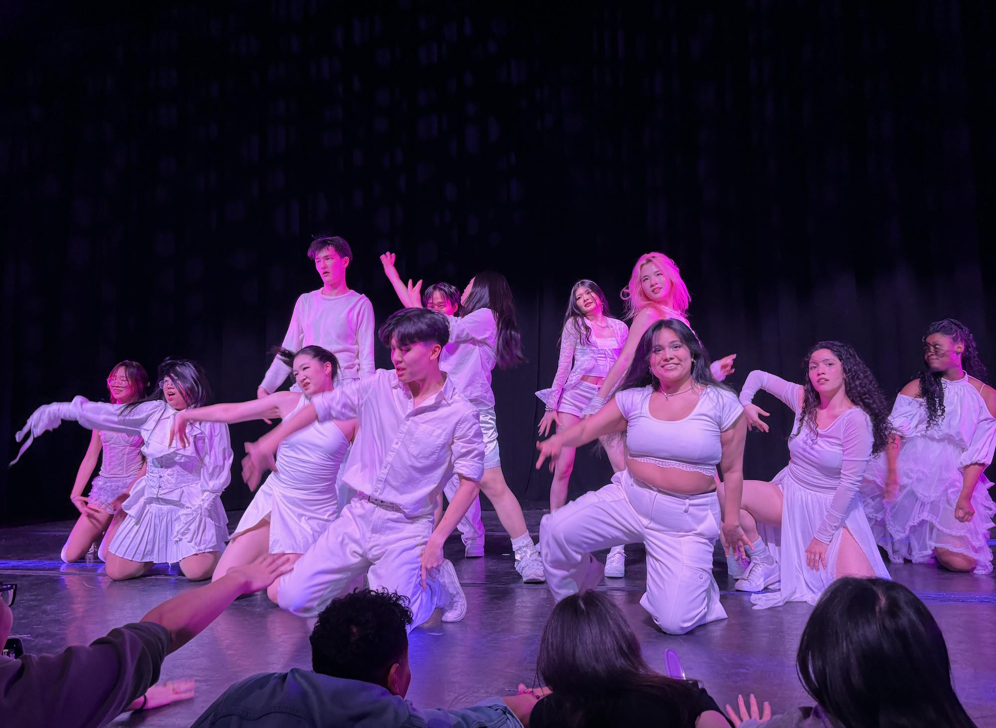 Several dancers in all white outfits dance on a stage with a black curtain in the background.