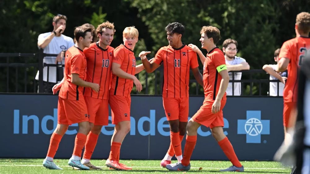 Five men in orange uniforms with black stripes put their arms around each other on a green soccer field.