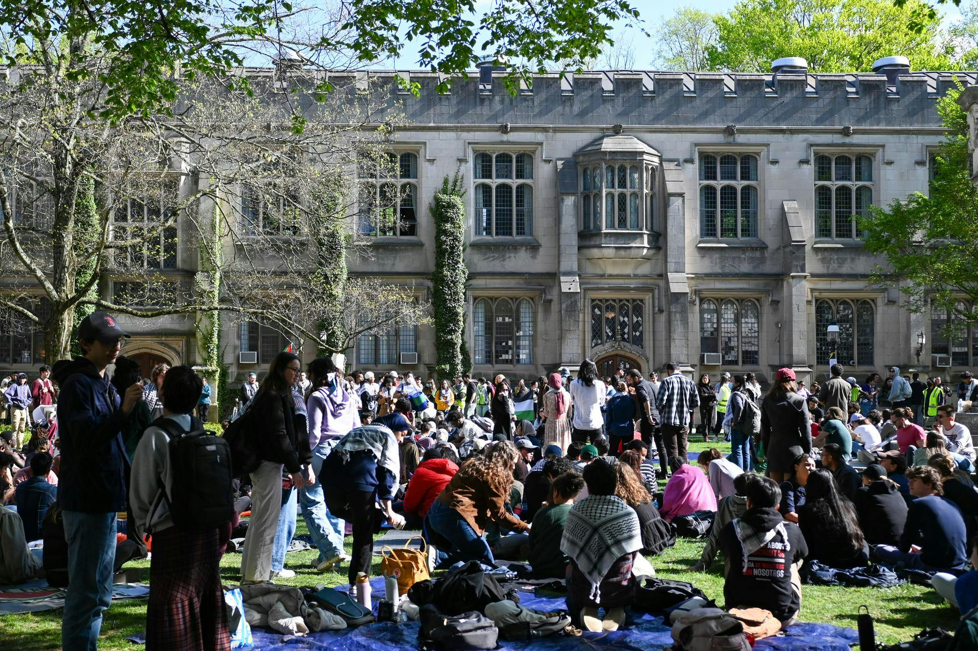 Students gather on a lawn in front of a Tudor Gothic style building on a sunny day. Most students are sitting on a picnic blankets with a few standing. 