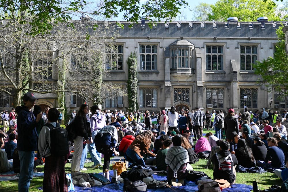 Students gather on a lawn in front of a Tudor Gothic style building on a sunny day. Most students are sitting on a picnic blankets with a few standing. 